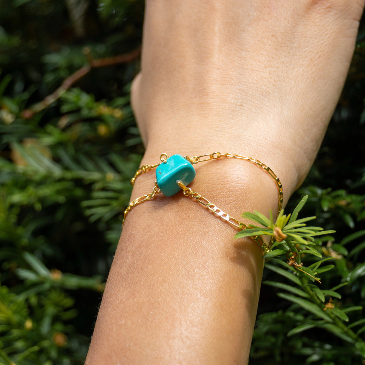 Gold bracelet with a turquoise stone on a wrist against a green leafy background