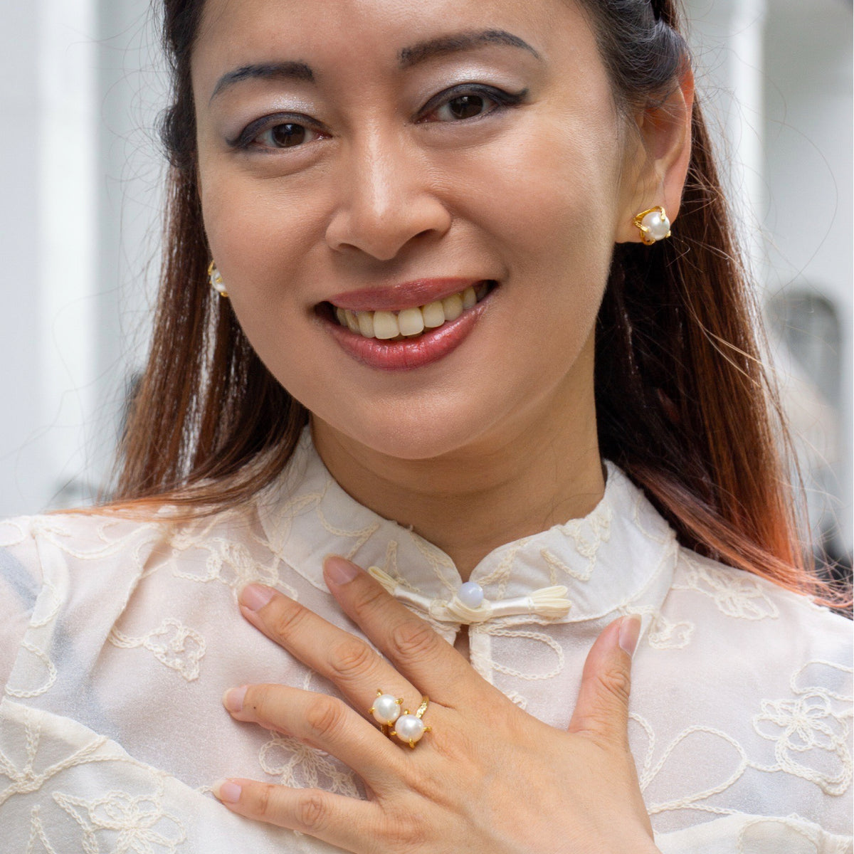 Woman wearing pearl earrings and ring, smiling outdoors.