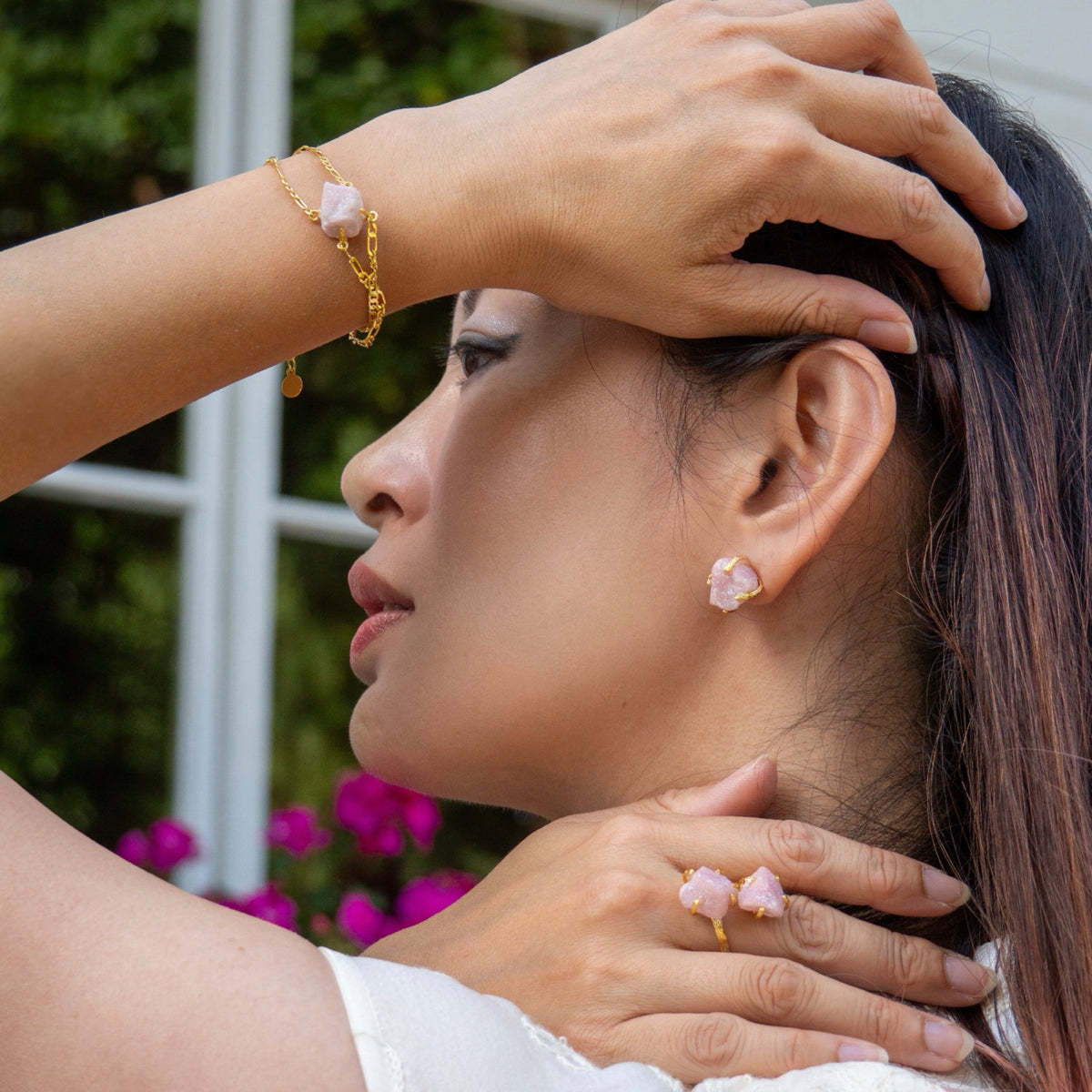 Woman wearing pink earrings and a ring, with a blurred background of greenery and flowers.