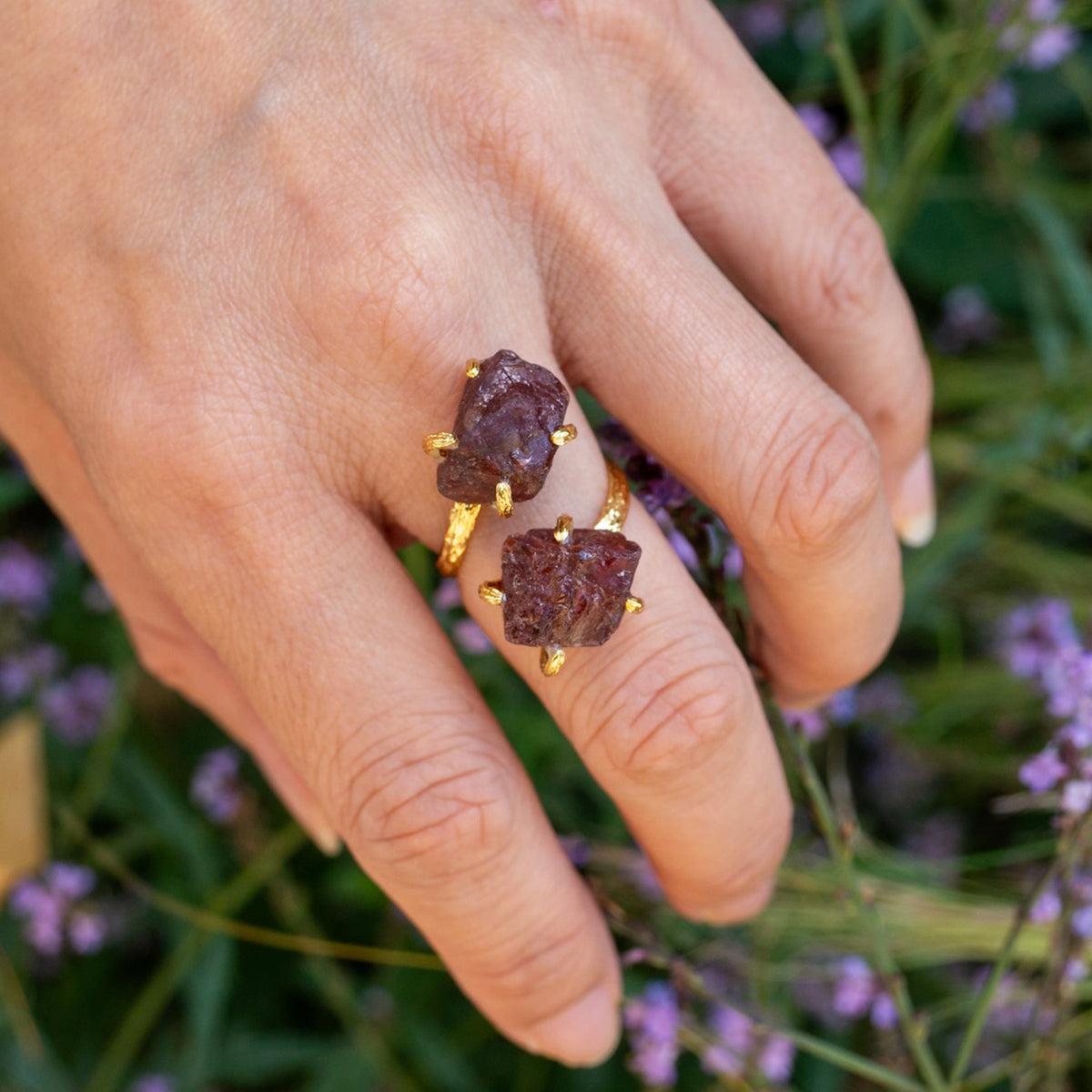 Hand wearing two purple gemstone rings with gold bands against a blurred natural background