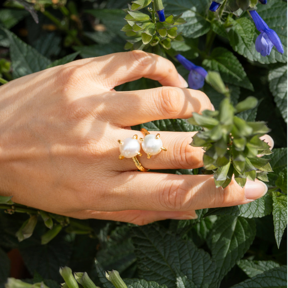Hand wearing a gold ring with pearl-like stones against a green leafy background