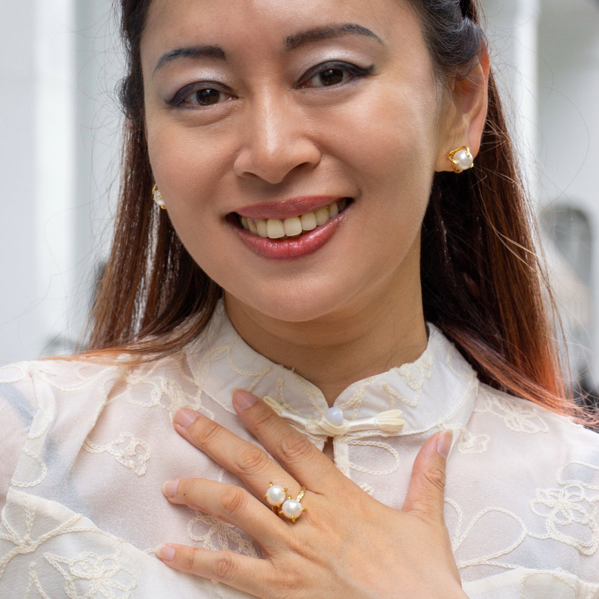 Woman wearing pearl earrings and rings, smiling outdoors.