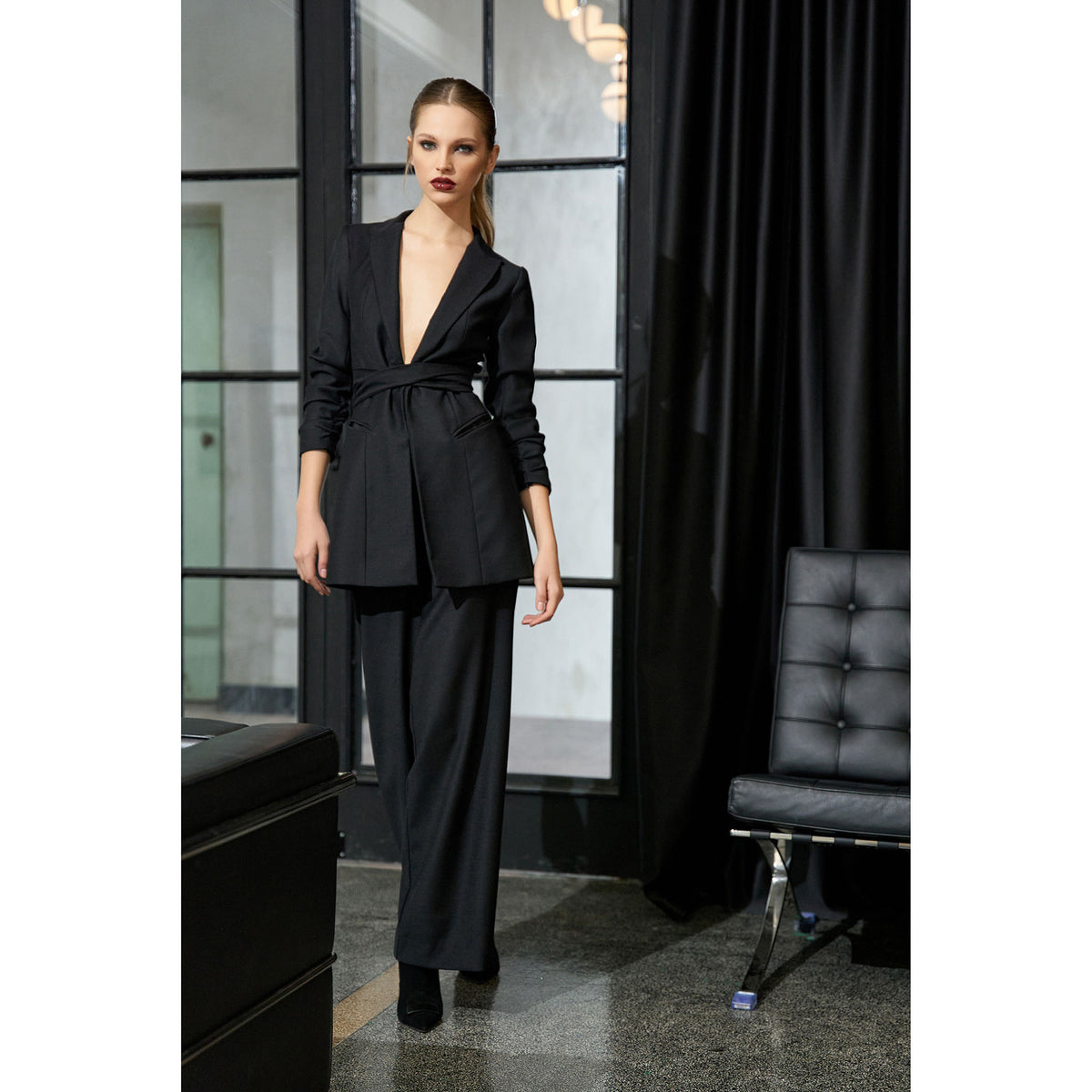 Woman wearing a black suit standing in an elegant room with a chair and window.
