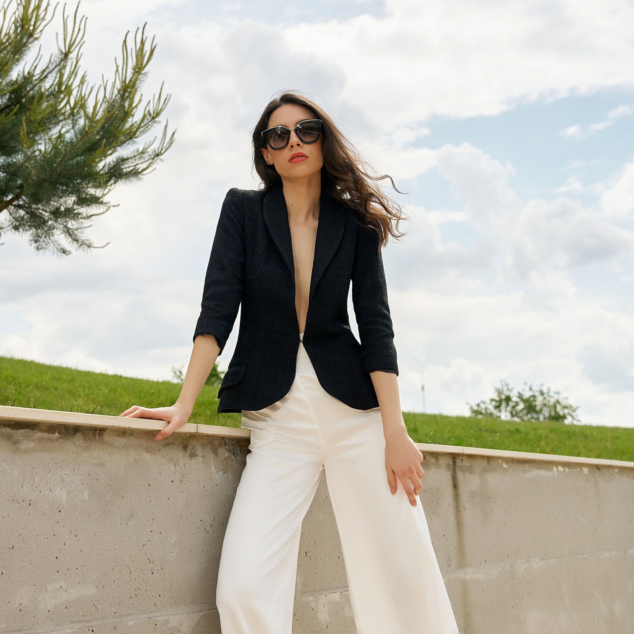 Woman in a black blazer and white pants standing outdoors with greenery and a cloudy sky in the background.