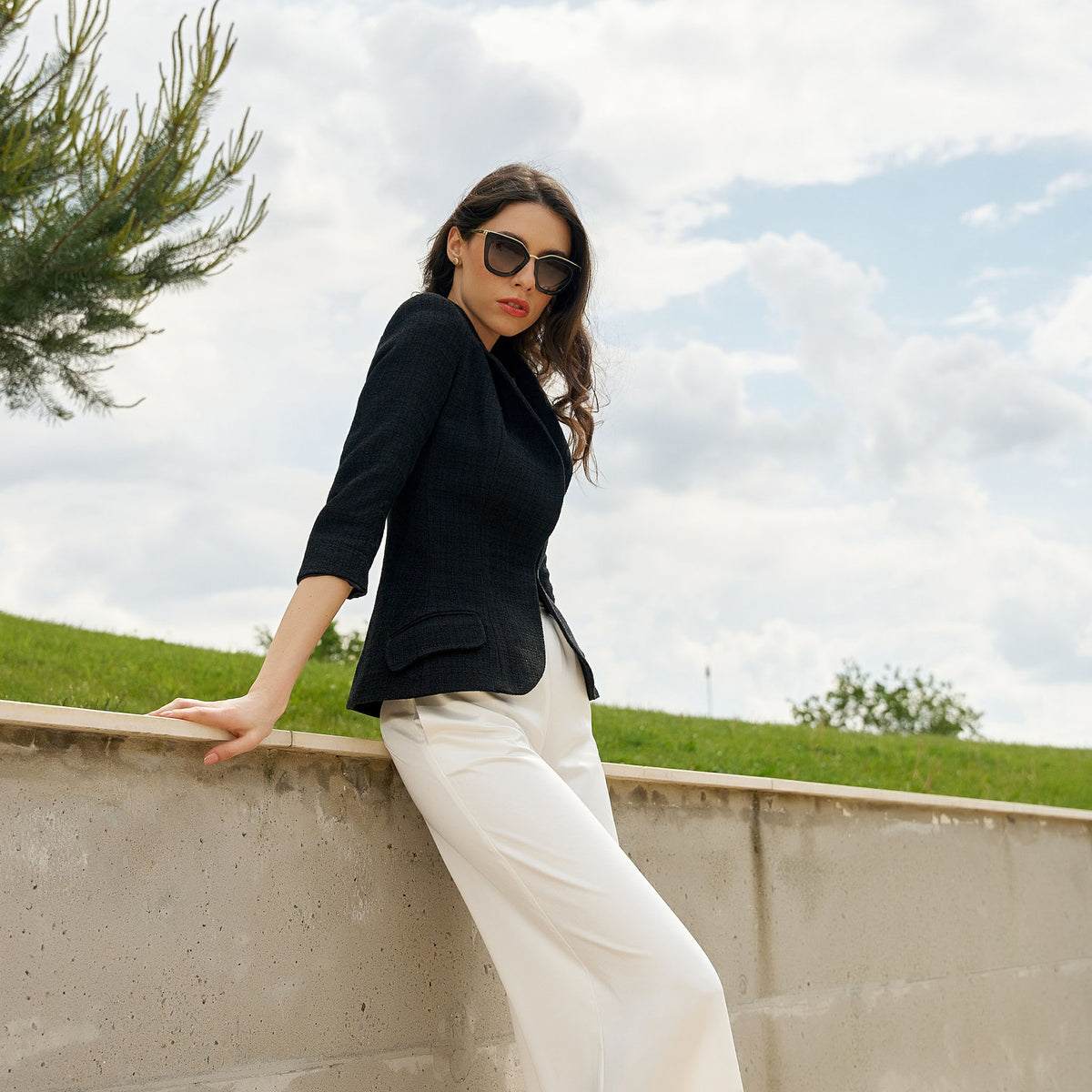 Woman in black jacket and white pants standing outdoors with a grassy hill and cloudy sky in the background.