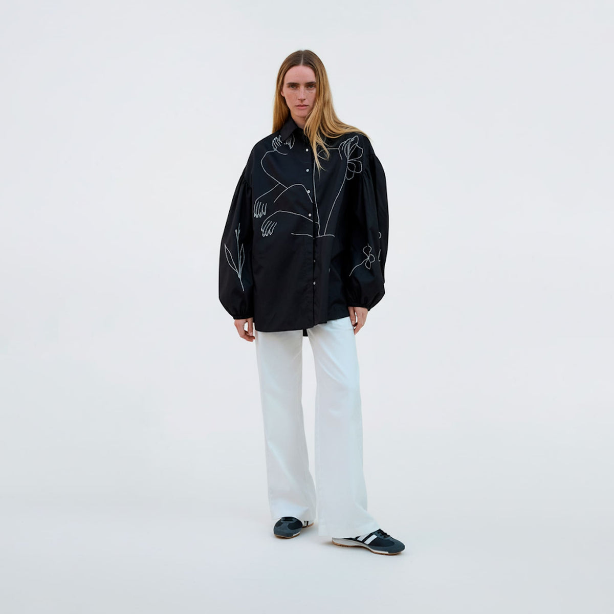 Person wearing a black embroidered jacket and white pants on a white background