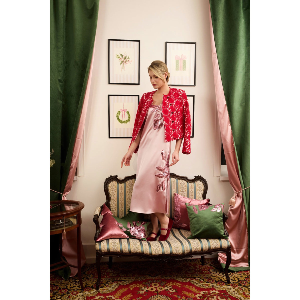 Woman in a pink dress and red jacket standing in a decorated room with green curtains and a patterned sofa.