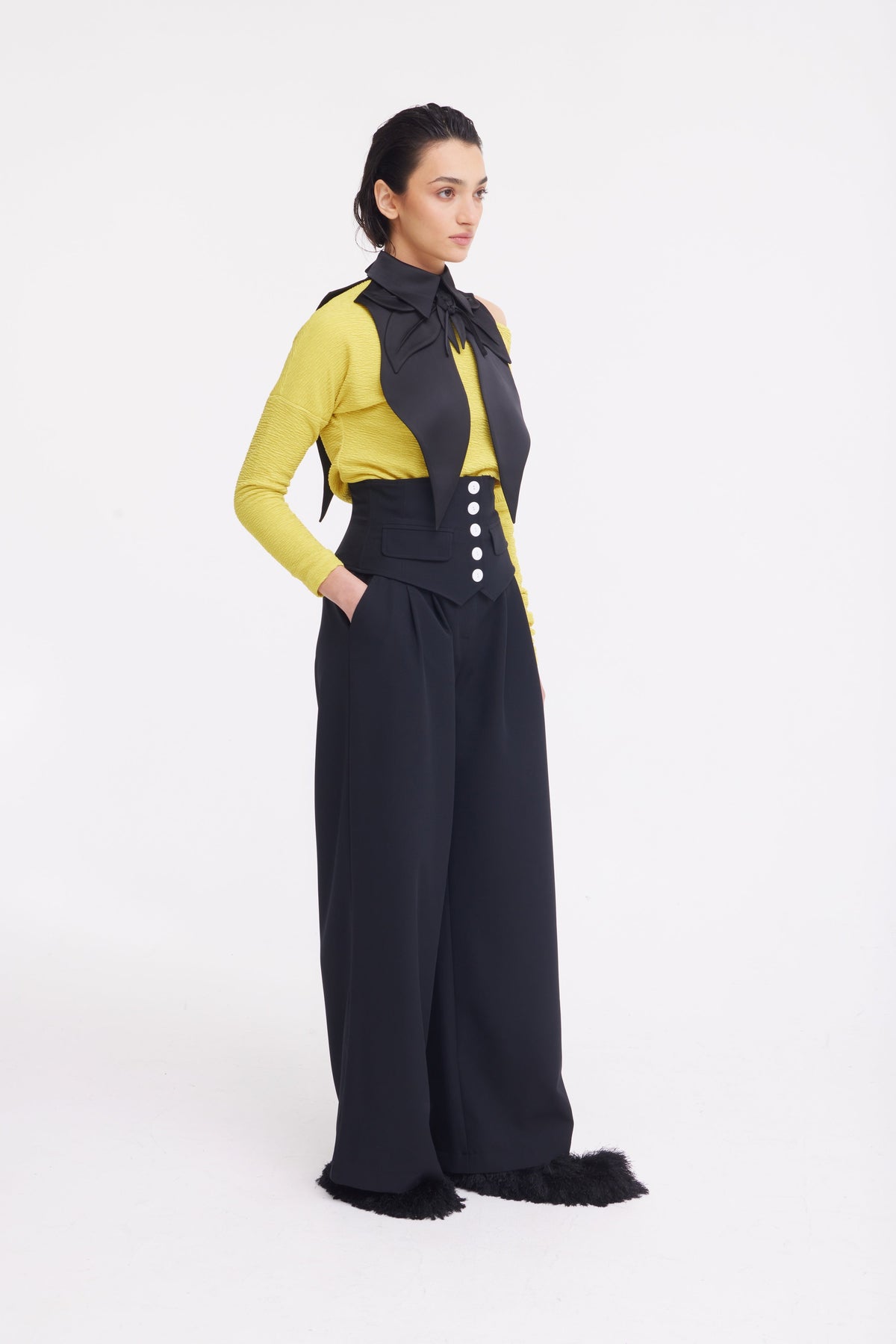 A woman stands against a white background wearing JULIA ALLERT's Buttoned Corset-Waist Black Tailored Trousers, detailed with white buttons and black furry accents at the hems.