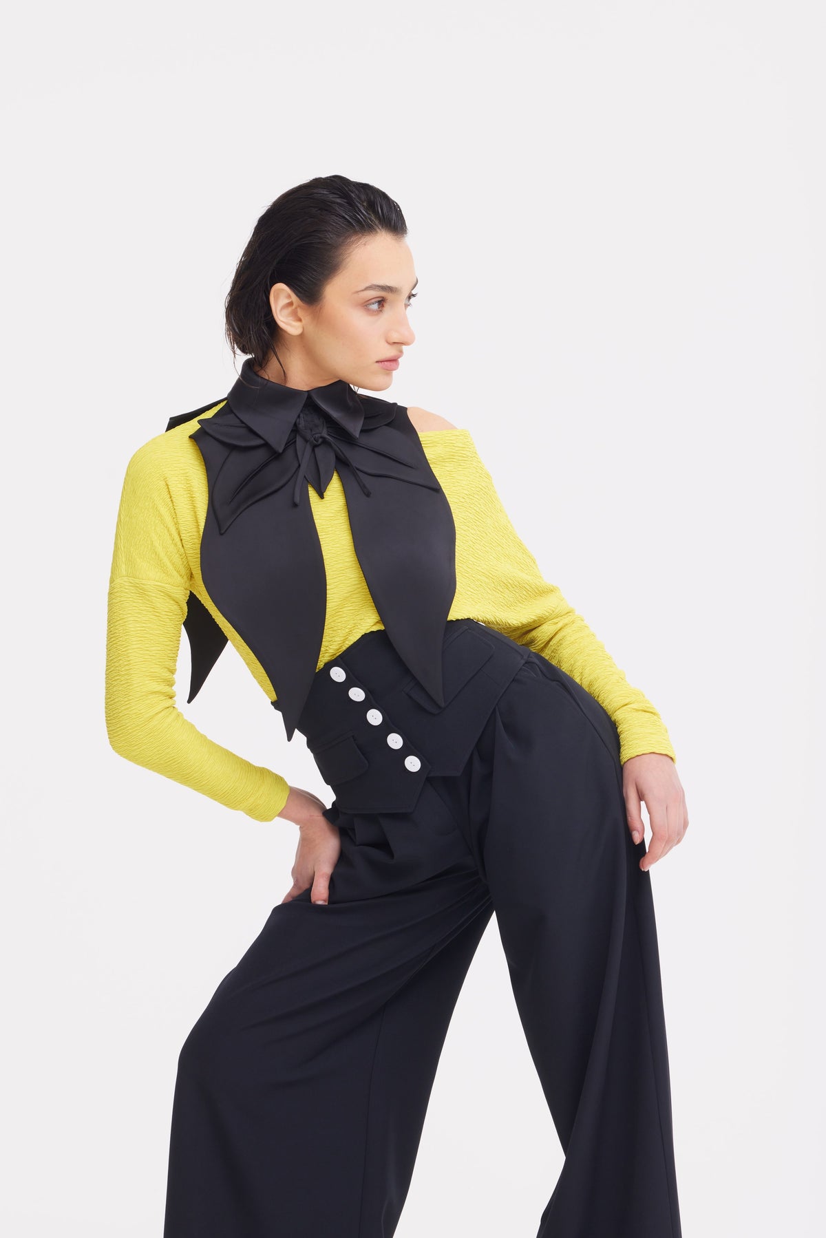 A woman poses against a white background wearing a yellow top, oversized black bow and collar, and JULIA ALLERT Buttoned Corset-Waist Black Tailored Trousers with white buttons; her dark hair is slicked back.