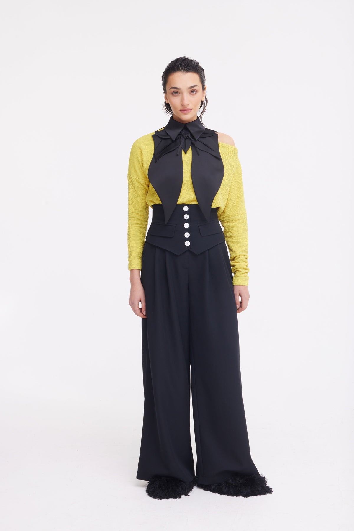 A woman stands against a white background, wearing a yellow long-sleeve top, black oversized bow tie, and JULIA ALLERT Buttoned Corset-Waist Black Tailored Trousers. She looks directly at the camera.