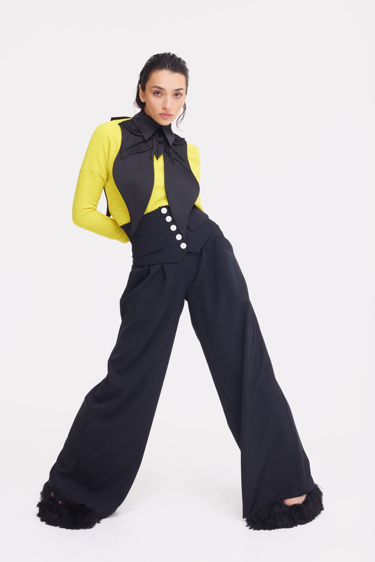 A woman poses confidently in JULIA ALLERT Buttoned Corset-Waist Black Tailored Trousers, paired with a yellow long-sleeve top, black vest with large buttons, and a black neck bow against a white background.