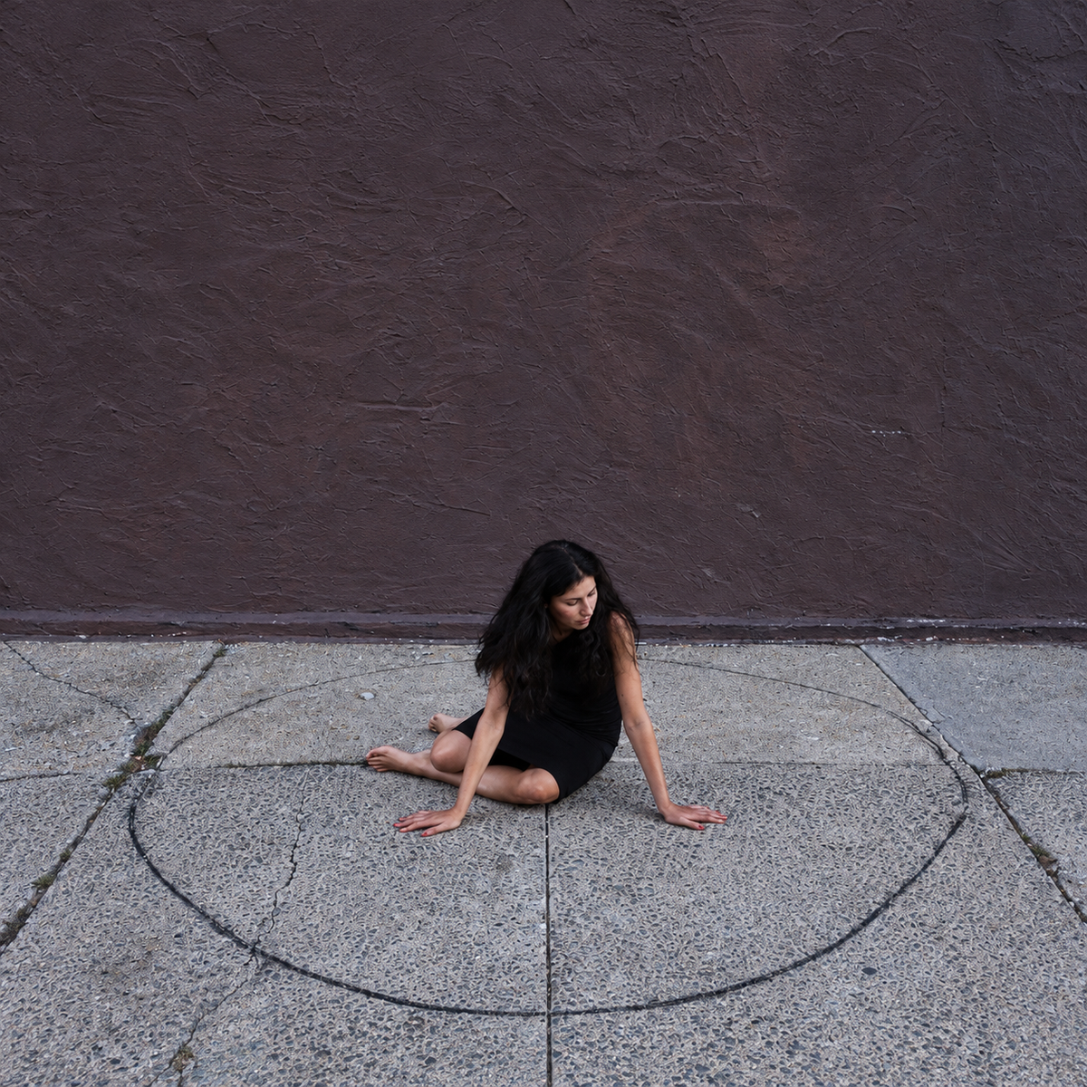 ALSU SHARM Circle II, fine art photography of a woman in a black dress sitting inside a drawn circle on a sidewalk in front of a brown wall, limited edition, buy contemporary art at DOORS NYC