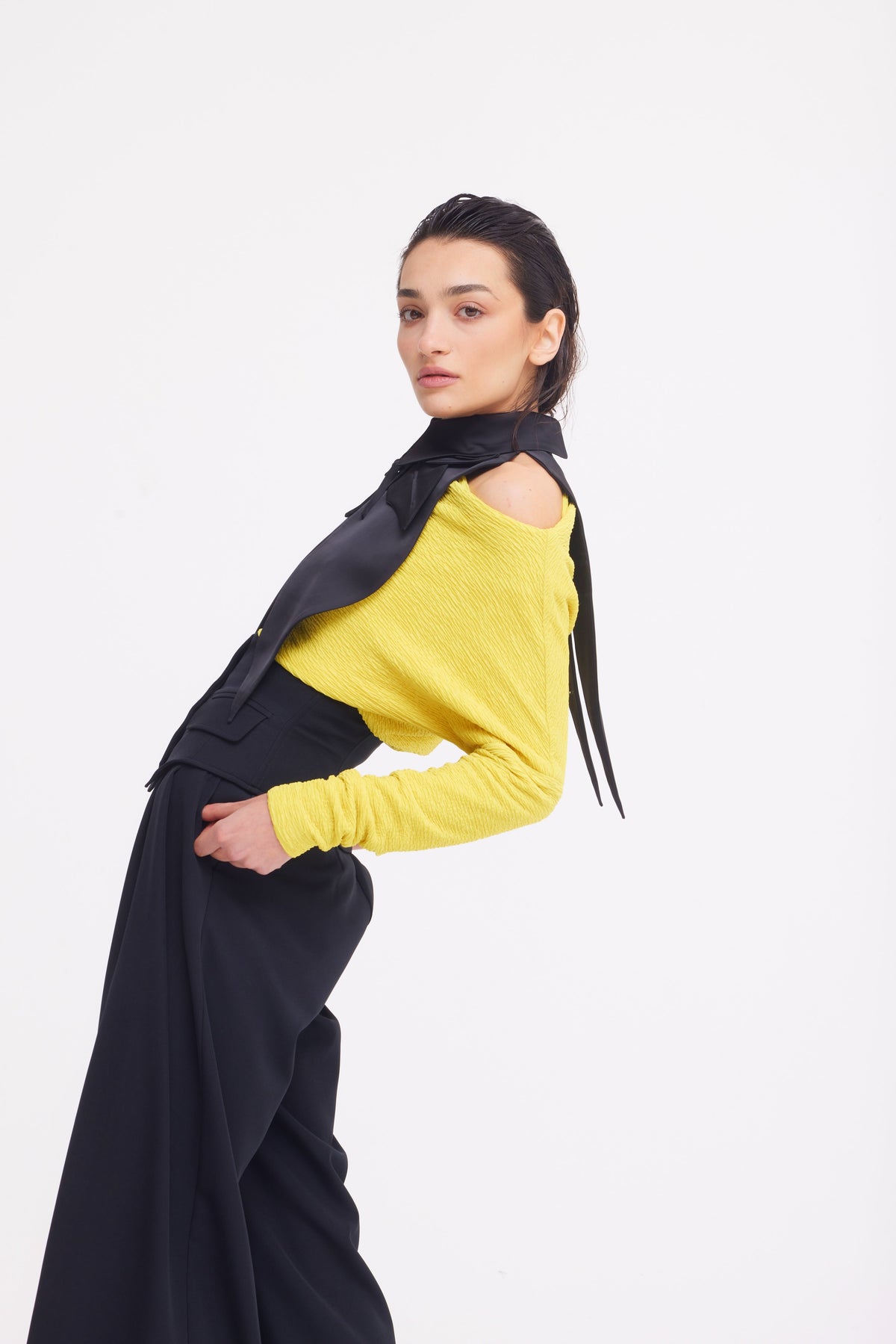 A woman poses against a white background in a yellow long-sleeve top with a shoulder cut-out and JULIA ALLERT Buttoned Corset-Waist Black Tailored Trousers, finished with a black neck scarf. She leans back, looking confidently at the camera.