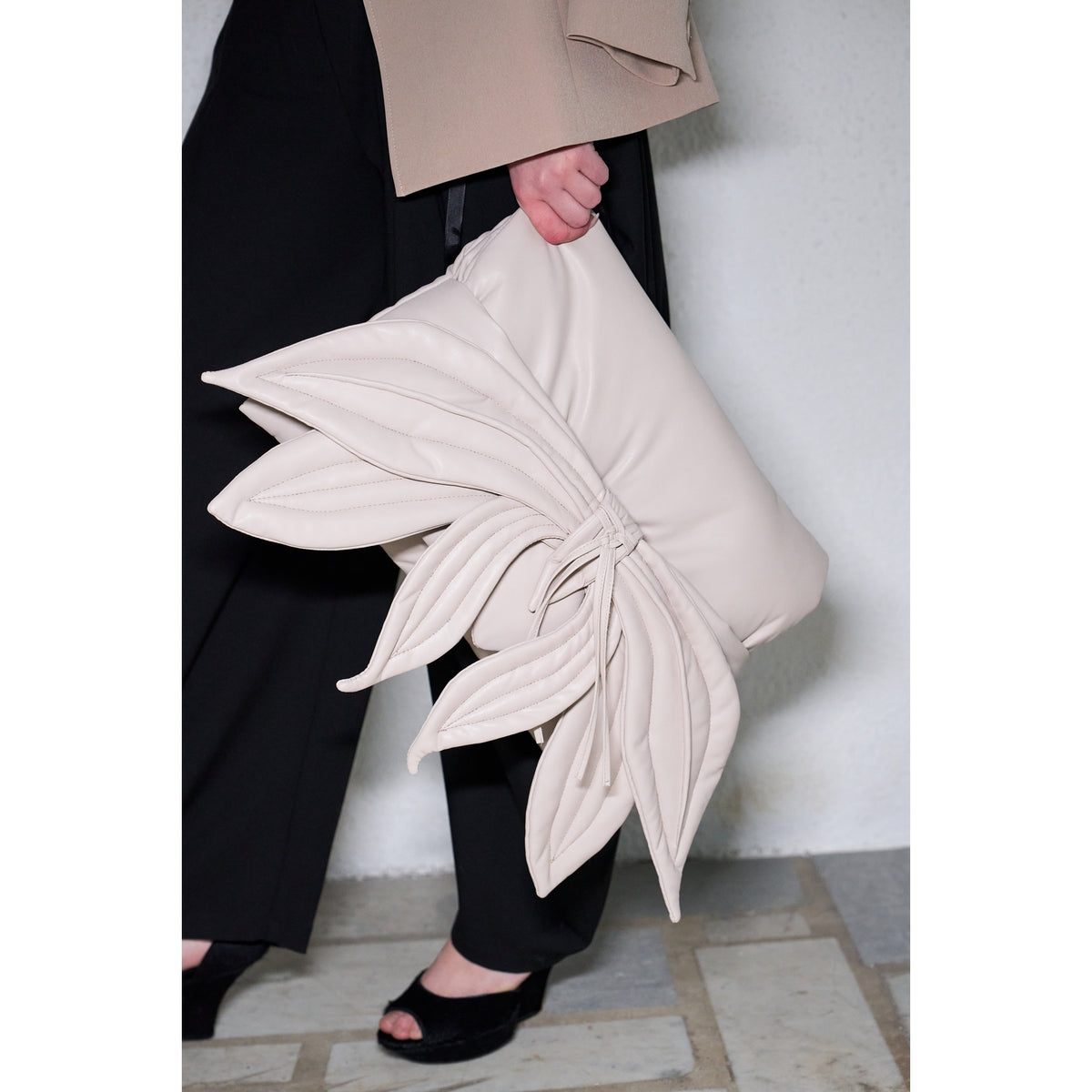 A person in black pants and open-toed black shoes holds the JULIA ALLERT Petal Faux Leather Evening Clutch Bag in ecru, featuring layered leaf-shaped details. A white wall and stone tile floor add modern flair to the look.