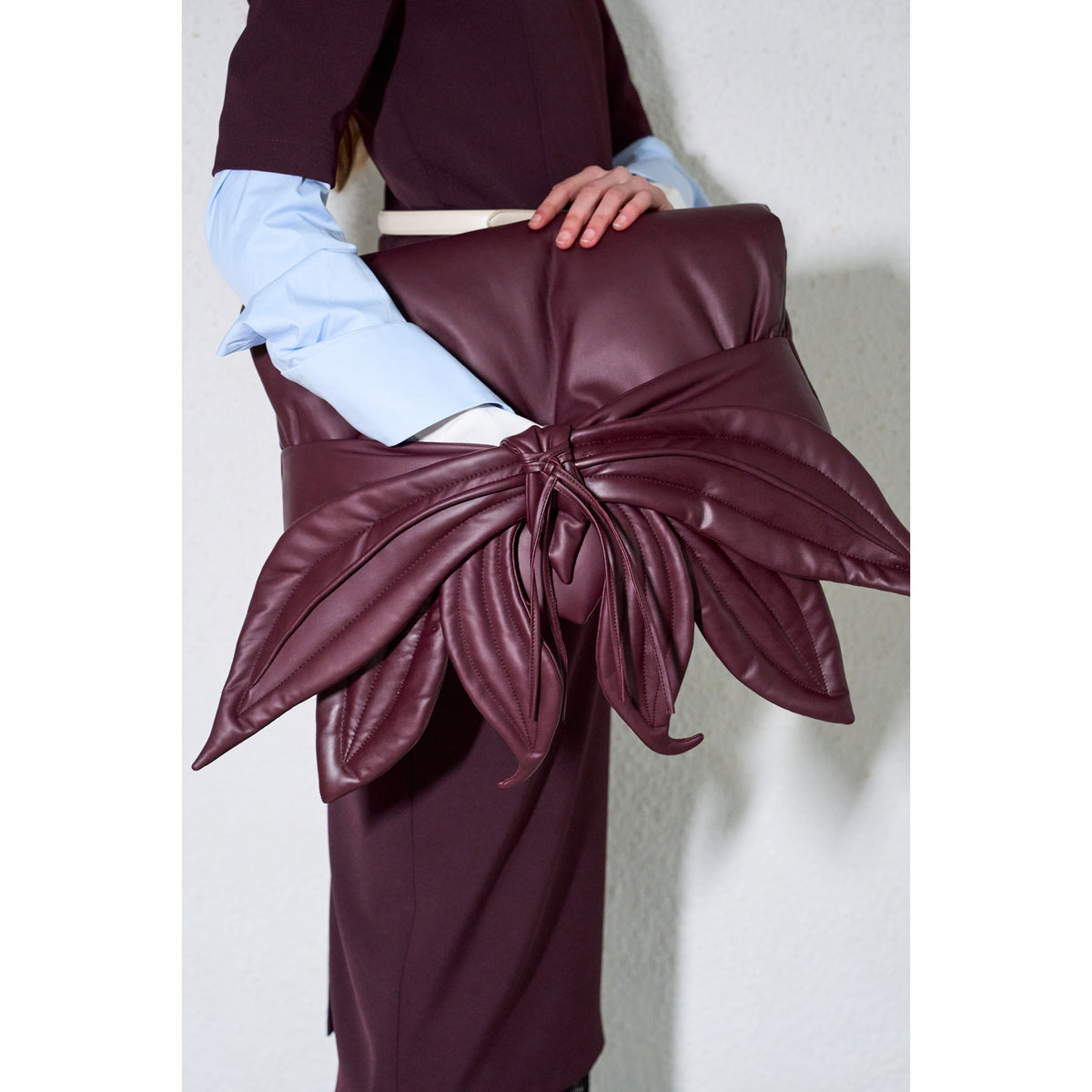 A person wearing a dark burgundy outfit with light blue sleeves holds the JULIA ALLERT Petal Faux Leather Evening Clutch Bag - Burgundy, a large flower-shaped clutch with oversized petals, against a plain white wall.