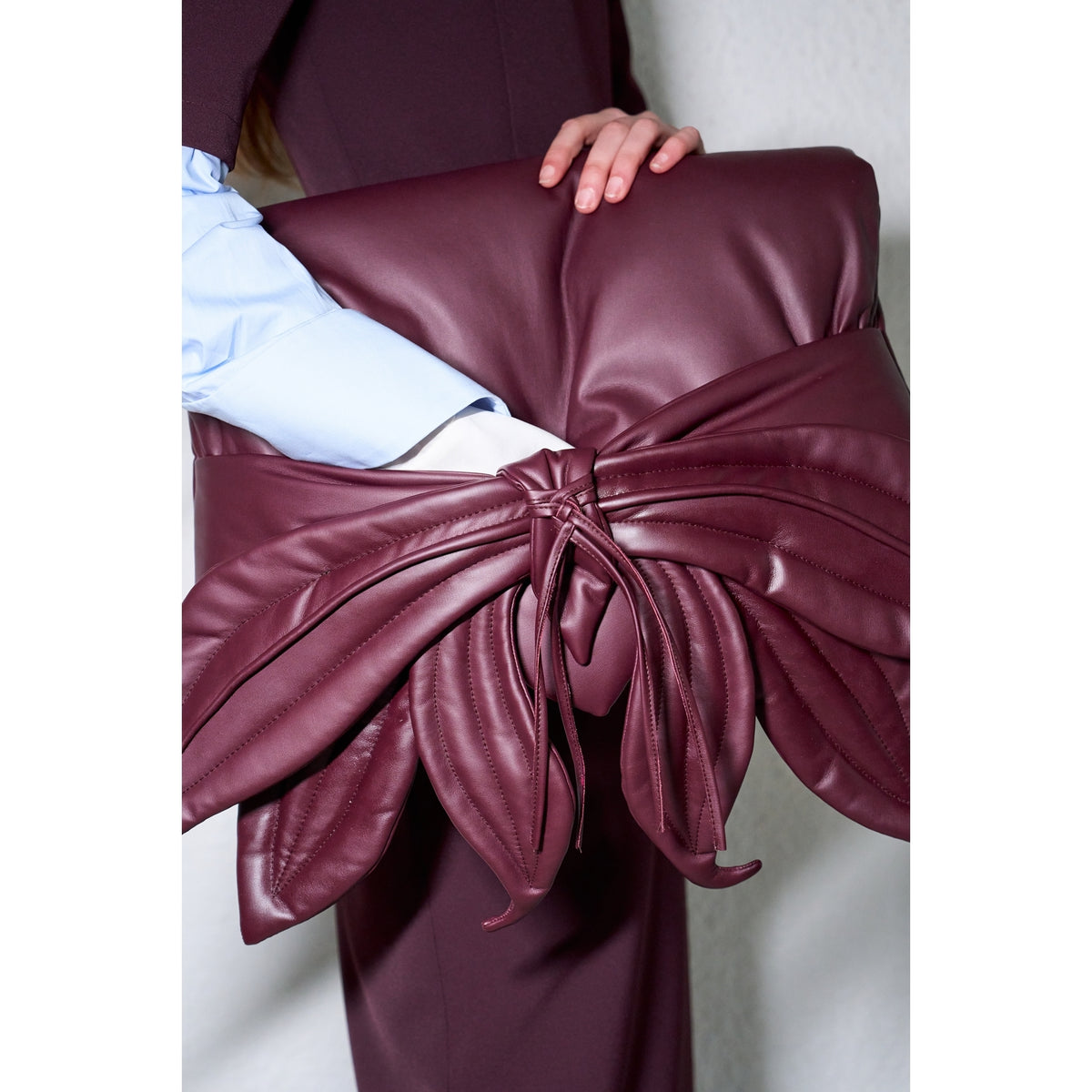 A person holds the JULIA ALLERT Petal Faux Leather Evening Clutch Bag - Burgundy, adorned with a leaf design and tie detail, stylishly paired with a pale blue shirt and dark maroon pants.