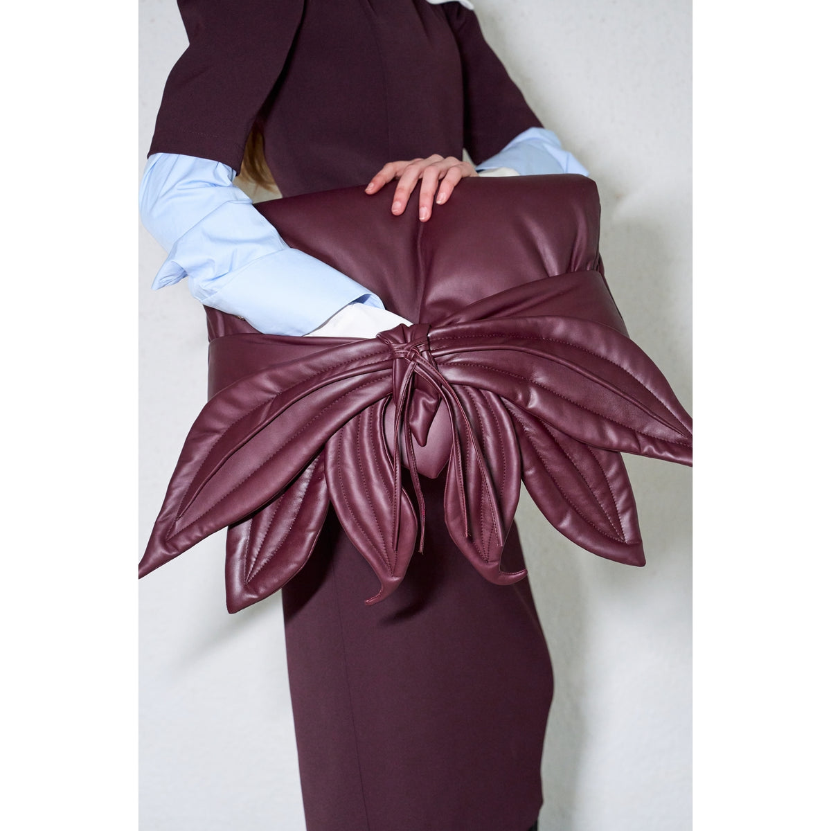 A person in a dark outfit holds the JULIA ALLERT Petal Faux Leather Evening Clutch Bag - Burgundy, a large clutch with decorative leather leaves, tucked under their arm and standing out against their light blue sleeves.