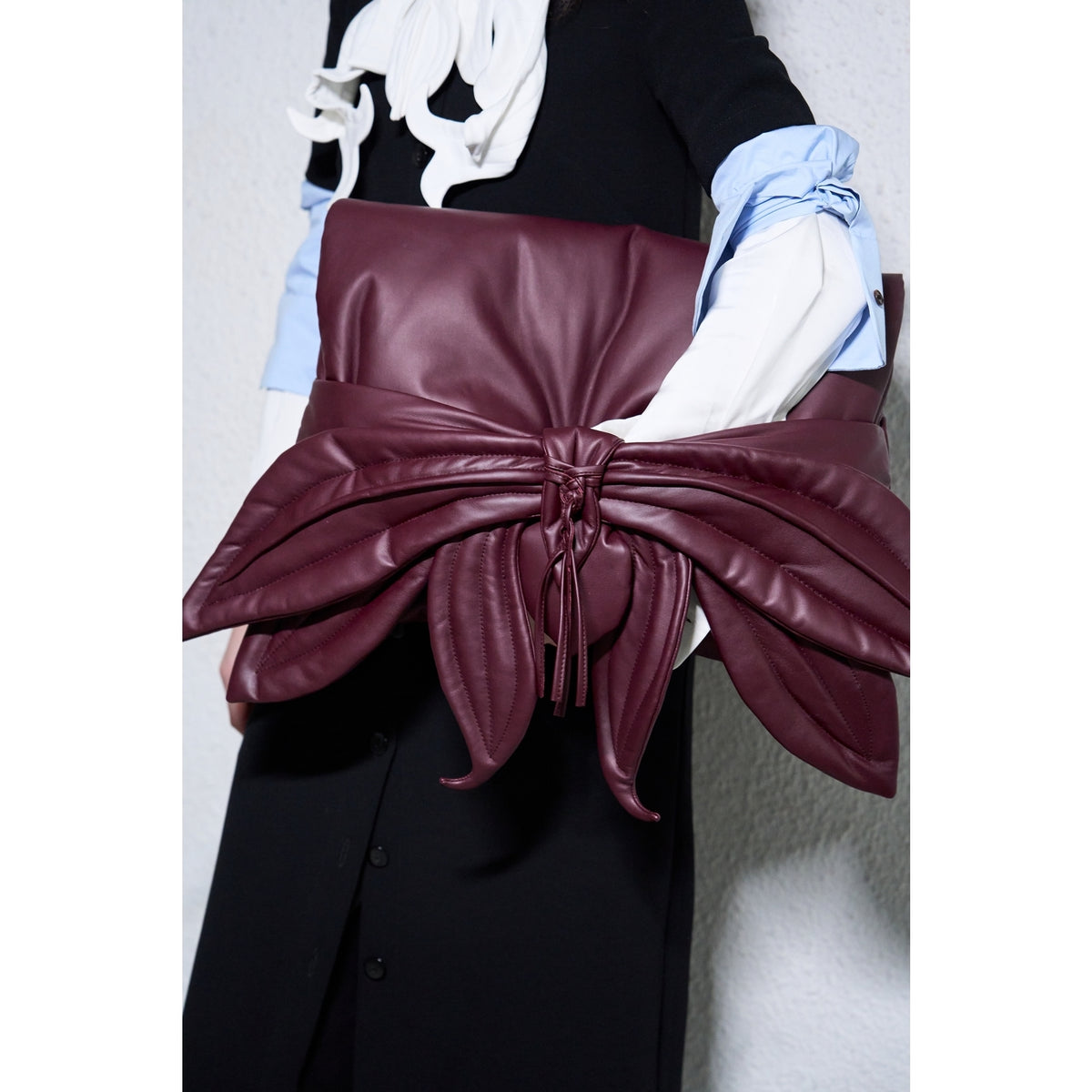 A person in a black and white outfit holds the JULIA ALLERT Petal Faux Leather Evening Clutch Bag - Burgundy, an eye-catching bow-shaped clutch with petal-like details and tasseled accents.