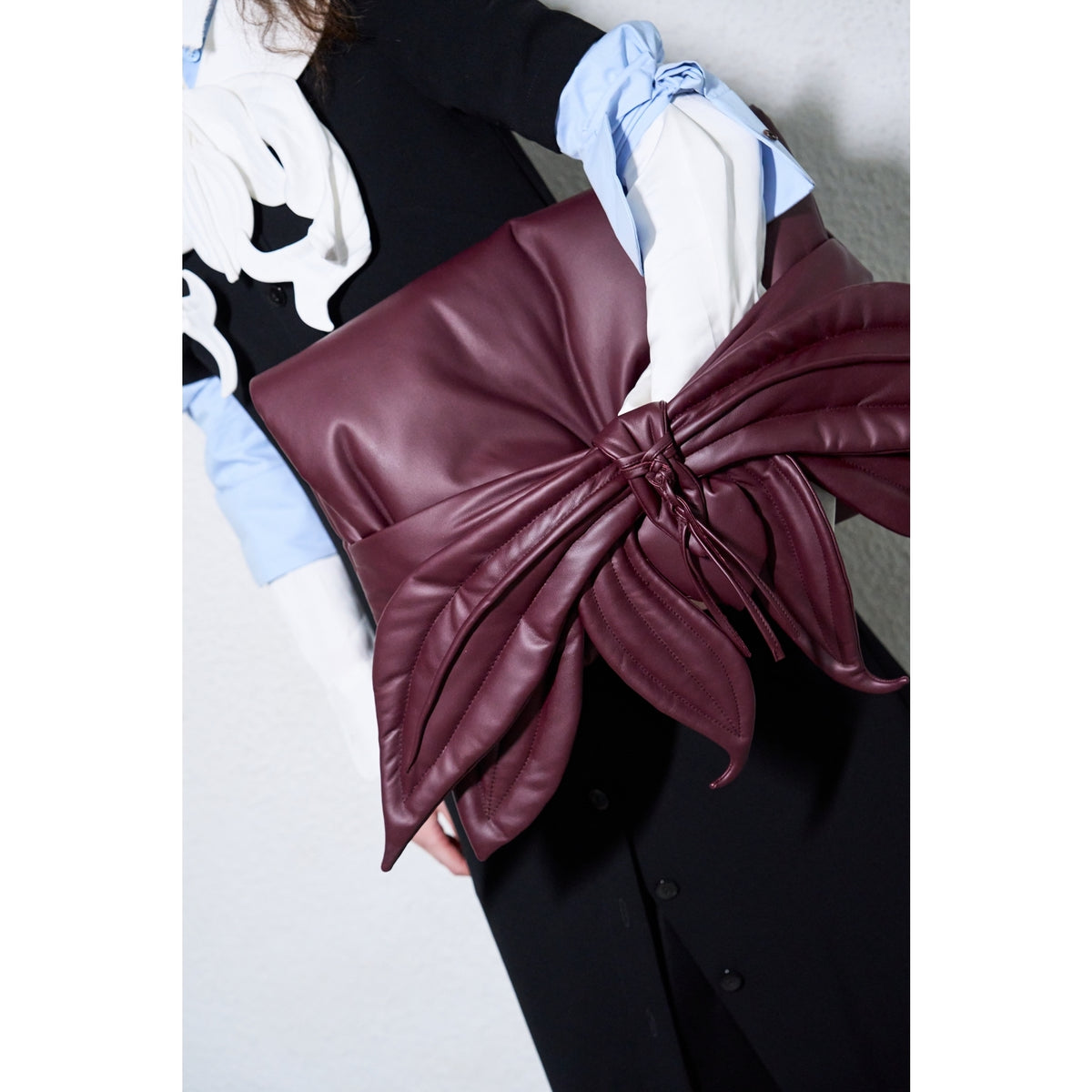 A person in a stylish outfit holds the JULIA ALLERT Petal Faux Leather Evening Clutch Bag in Burgundy, featuring an oversized bow. They wear a shirt with blue and white sleeves and a black vest or dress with white abstract details.