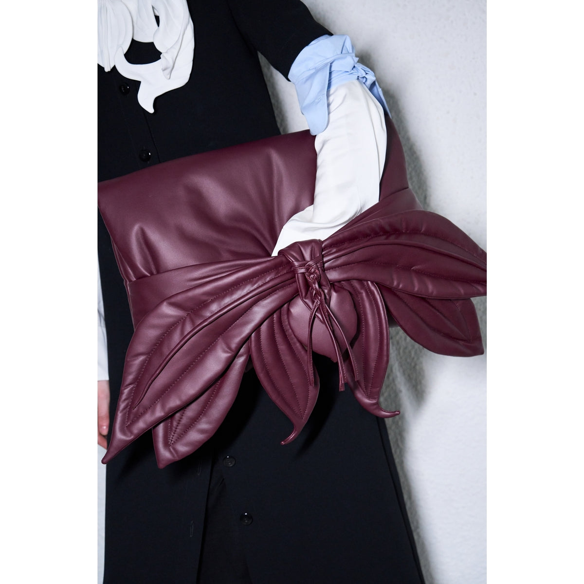A person holds the JULIA ALLERT Petal Faux Leather Evening Clutch Bag in burgundy, a large clutch with a striking leaf-inspired bow, while dressed in a black dress, white ruffled collar, and white gloves with blue cuffs.