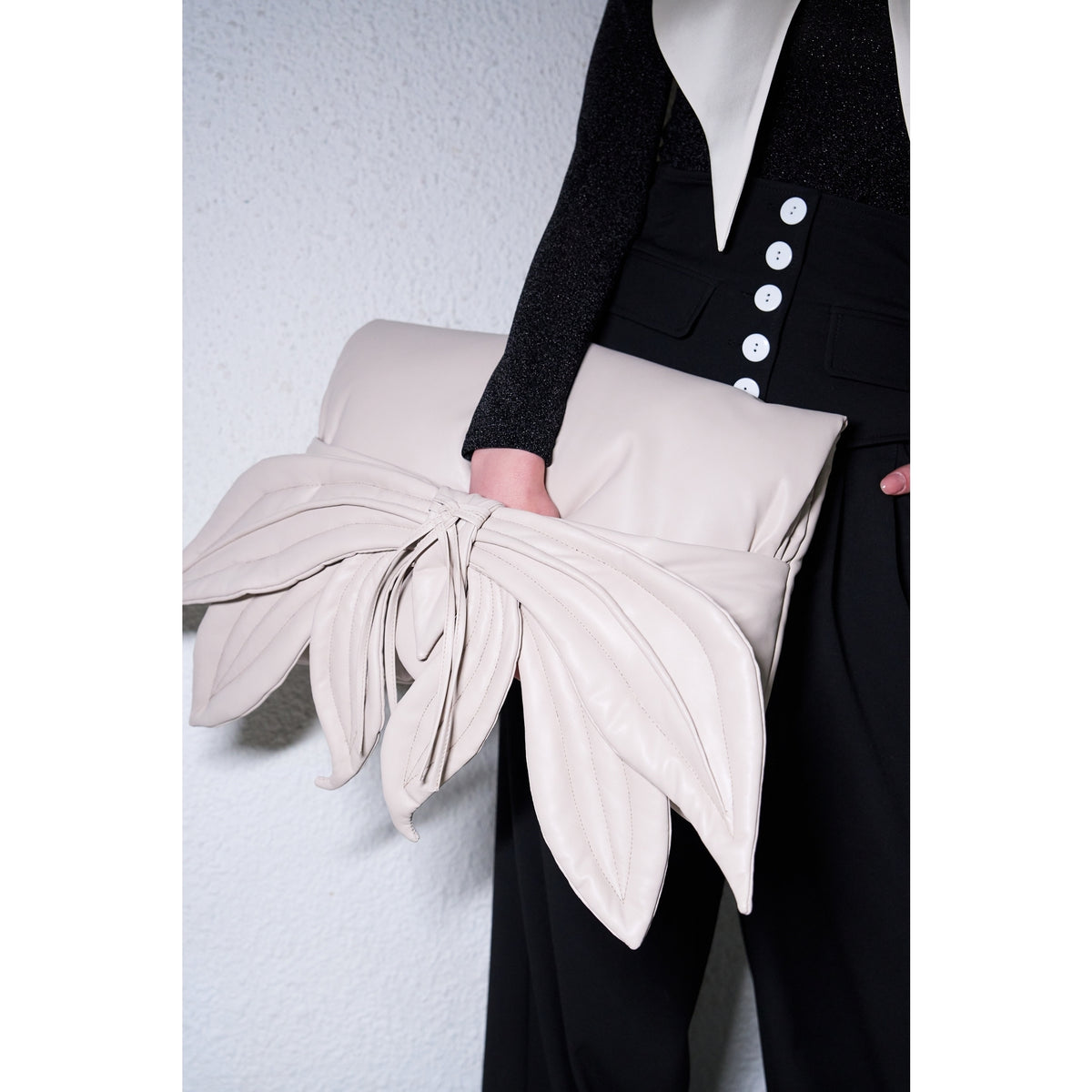Wearing black pants with white buttons and a black top, a person holds the JULIA ALLERT Petal Faux Leather Evening Clutch Bag in Ecru, featuring layered, petal-like details. A plain textured white wall serves as the background.