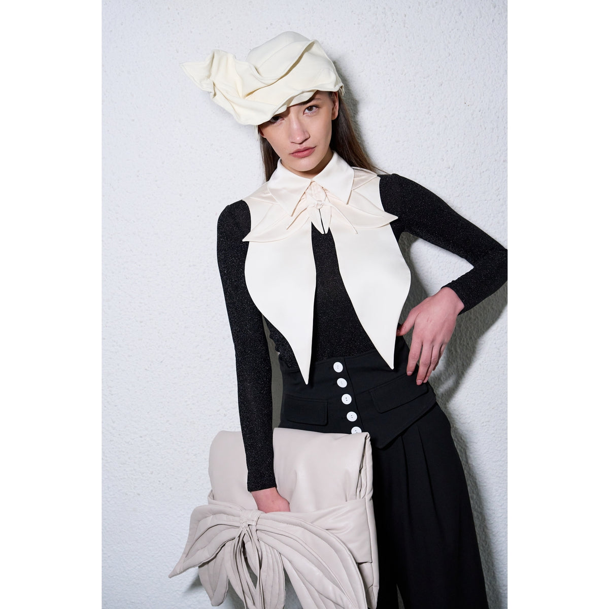 A woman poses against a white textured wall in JULIA ALLERT, wearing a black long-sleeve top, high-waisted pants with white buttons, a sculptural hat, and holding the Petal Faux Leather Evening Clutch Bag in ecru.