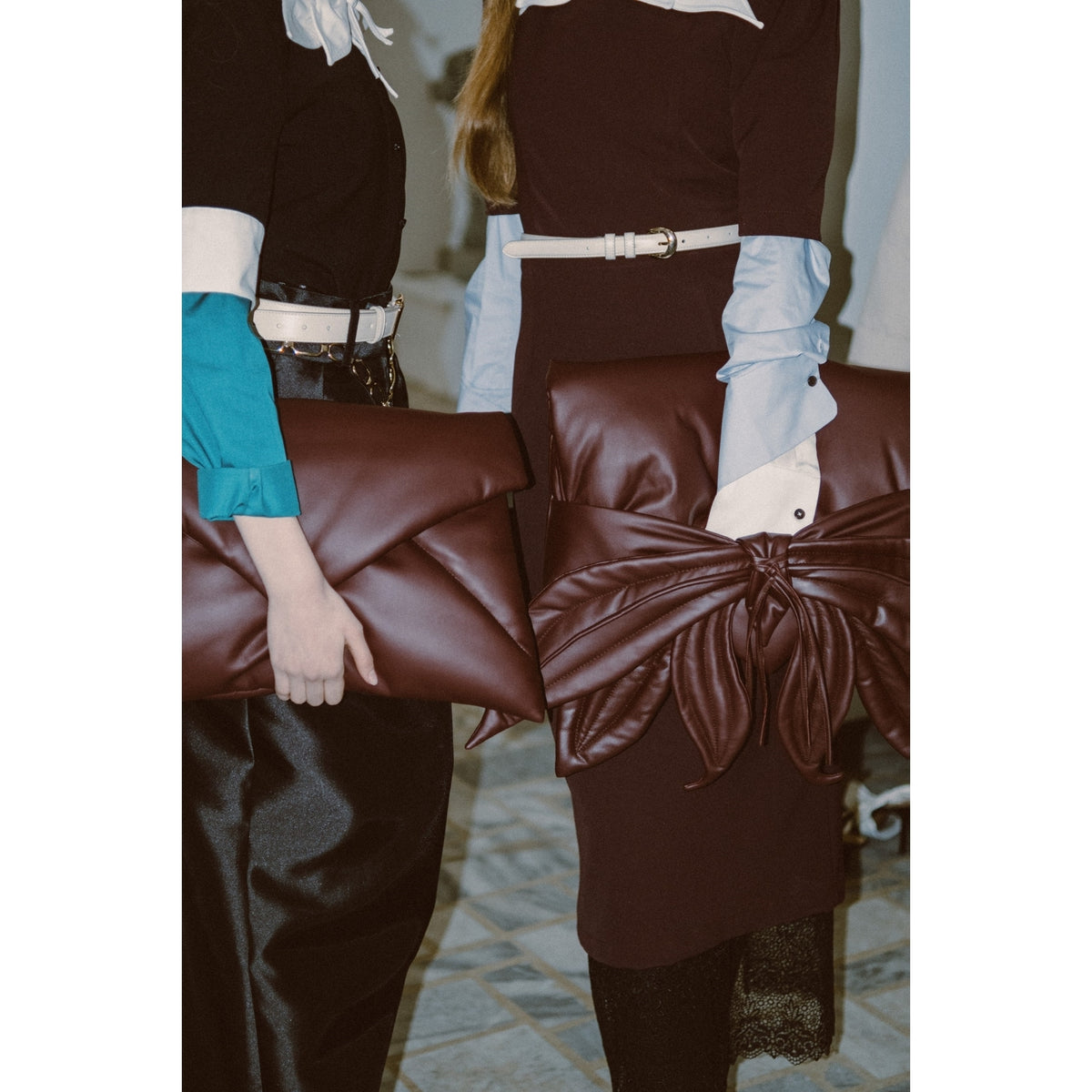 Two people stand side by side, each holding a JULIA ALLERT Petal Faux Leather Evening Clutch Bag in Burgundy. Wearing belted dark outfits with light blue and white sleeves, their faces are not visible in the frame.