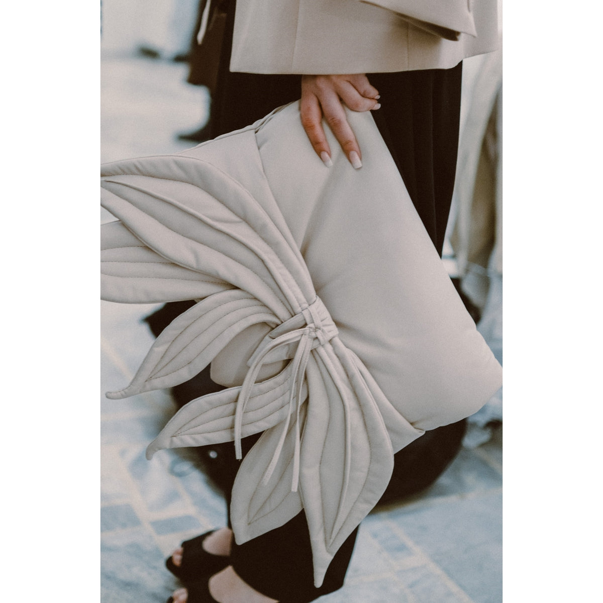 A person holds the JULIA ALLERT Petal Faux Leather Evening Clutch Bag in Ecru, featuring decorative petal-inspired details and a tied center, paired with black pants and open-toe shoes for a chic look.