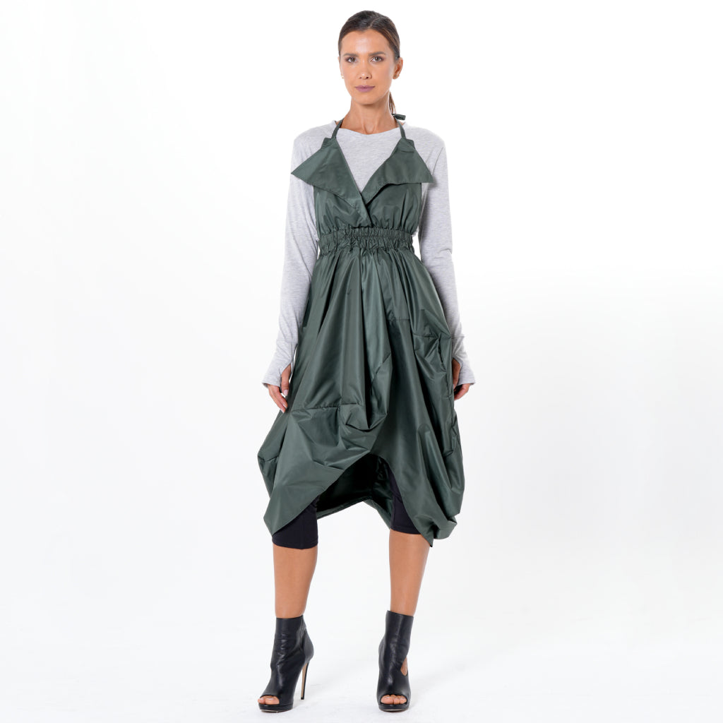 A woman faces forward against a white background, wearing the METAMORPHOZA Draped Balloon Pinafore Dress in Green over a long-sleeve gray top, black knee-length shorts, and black open-toe heeled ankle boots.