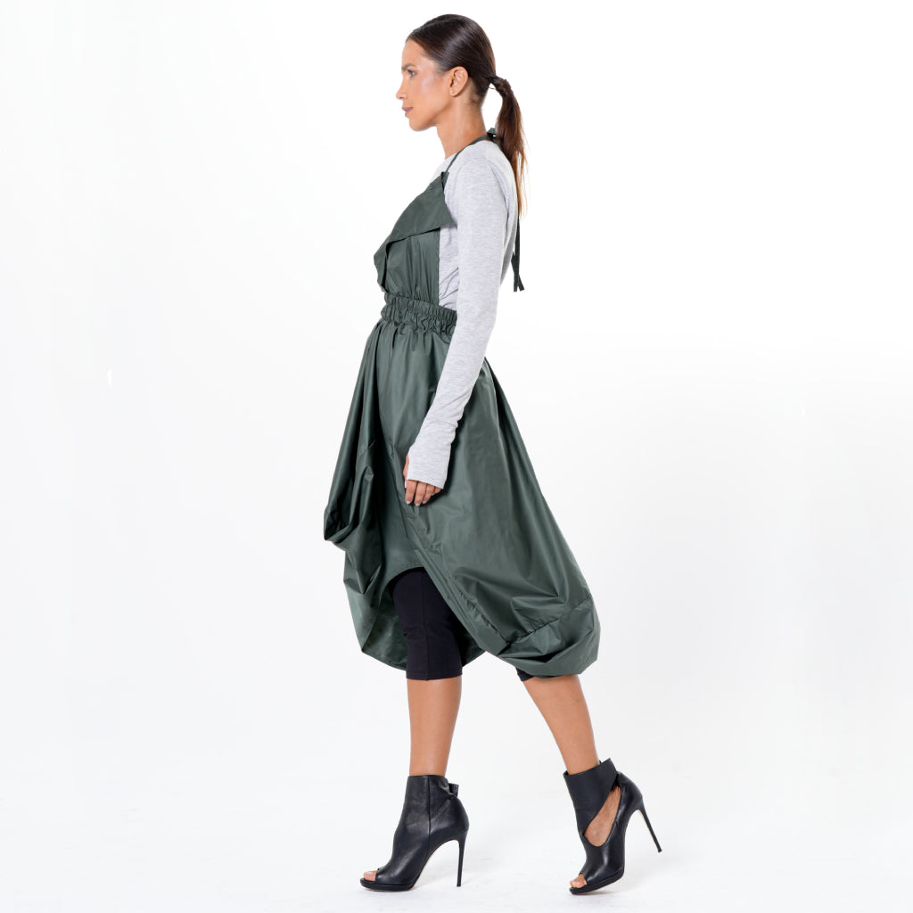 Woman in profile against a white background, wearing METAMORPHOZA&#39;s Draped Balloon Pinafore Dress in green over a gray long-sleeve top, paired with black leggings and black high-heeled open-toe ankle boots. Hair is styled in a ponytail.