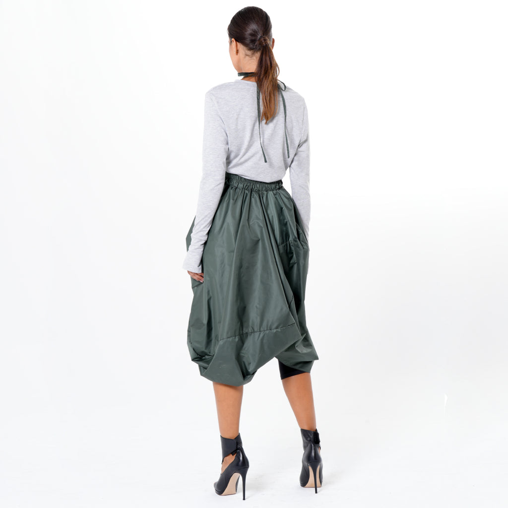 A woman with her hair tied back stands against a white background, wearing the METAMORPHOZA Draped Balloon Pinafore Dress in green over a gray long-sleeve top and dark high-heeled shoes.