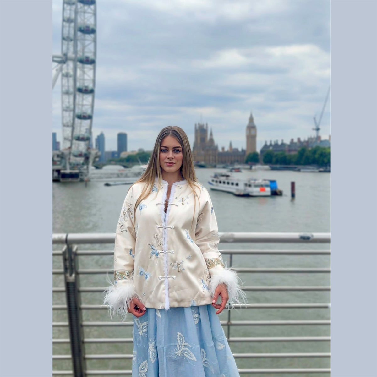 Woman in a white embroidered jacket with feathered sleeves standing in front of the London Eye and the Houses of Parliament.