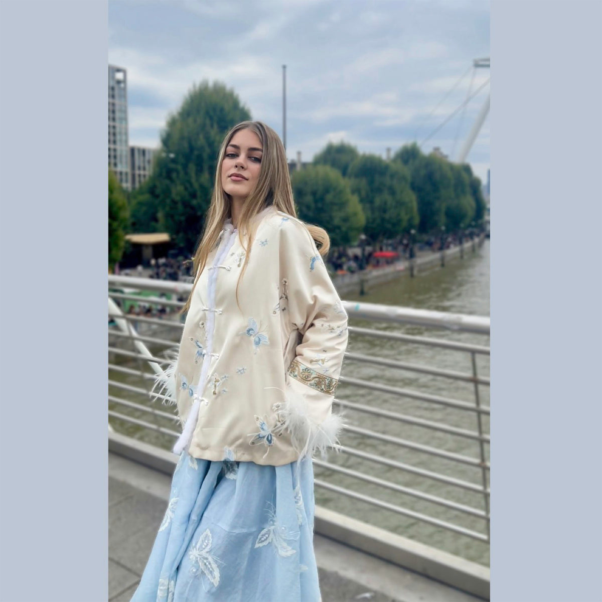 Woman wearing a light-colored embroidered jacket and light blue pants standing on a bridge with a cityscape background.