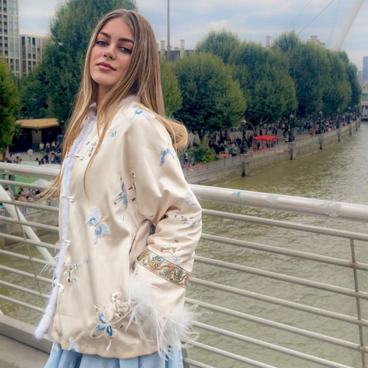 Woman in an embroidered jacket standing on a bridge by a river with trees and buildings in the background.