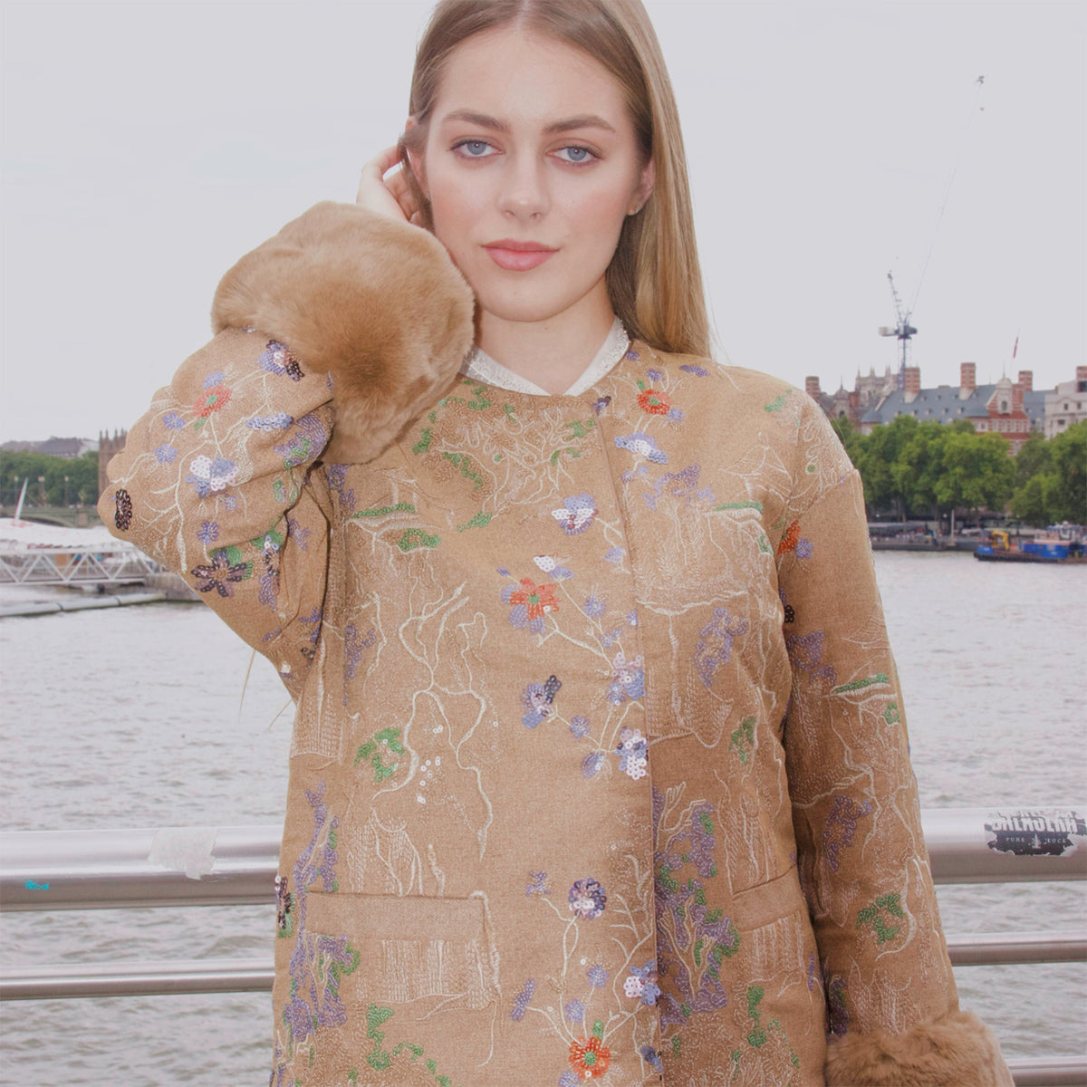 Woman wearing a floral embroidered coat with fur trim by a waterfront.