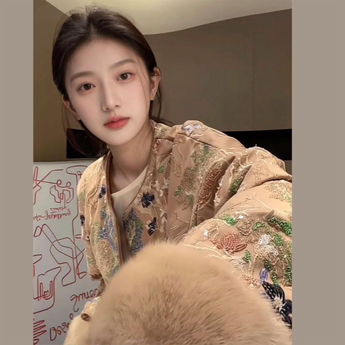 Person wearing a beige embroidered jacket in a room with a wall featuring abstract art.