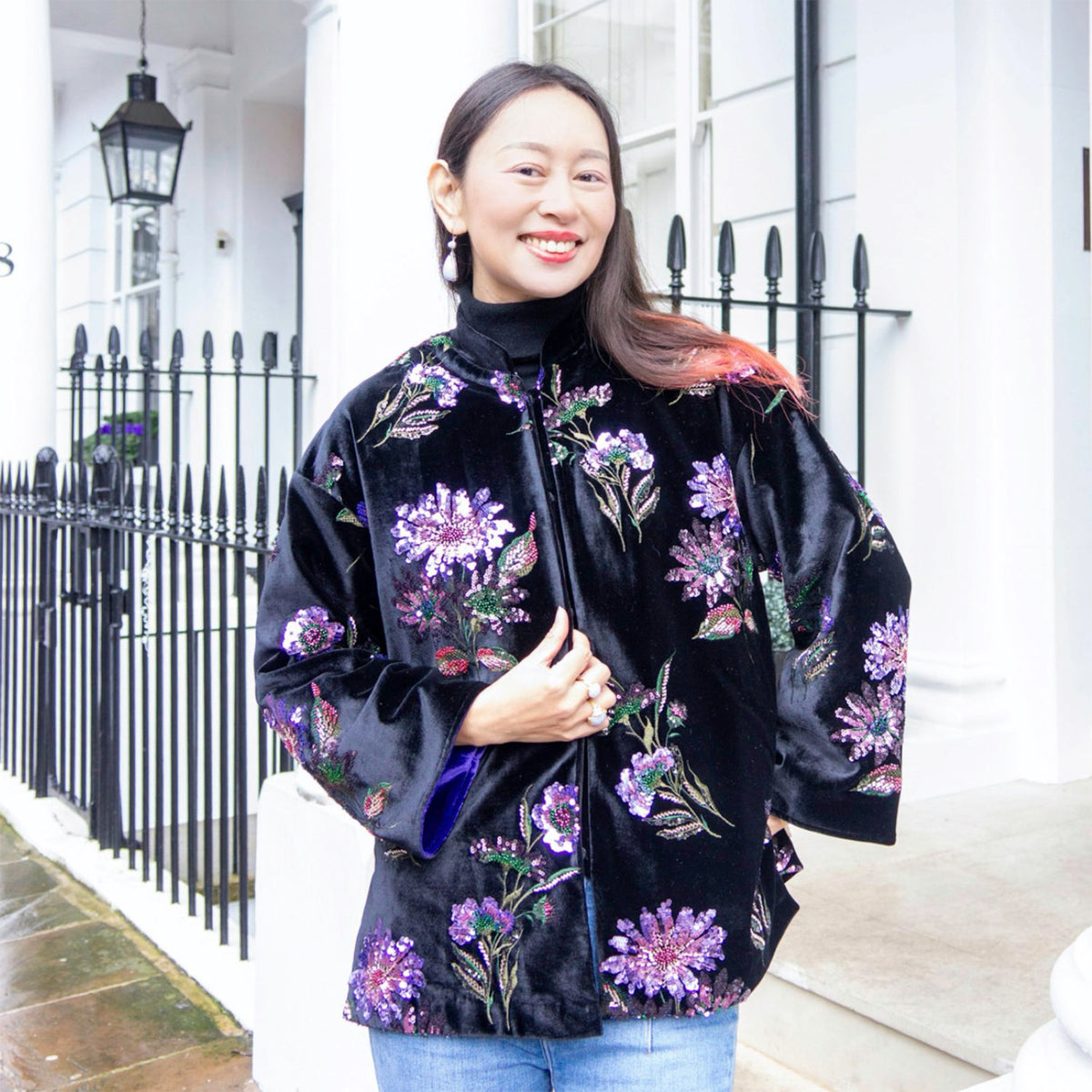 Woman wearing a black floral jacket standing in front of a white building with black railings.