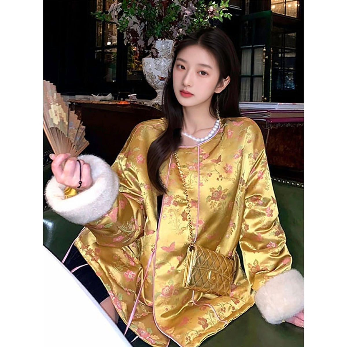 Woman in a traditional yellow outfit with fur cuffs holding a fan indoors.