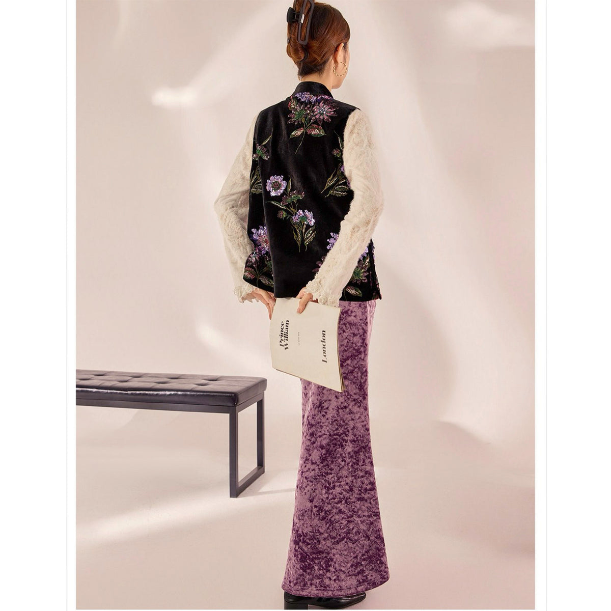 Person wearing a black floral vest over a white lace top with purple pants, holding a white bag.