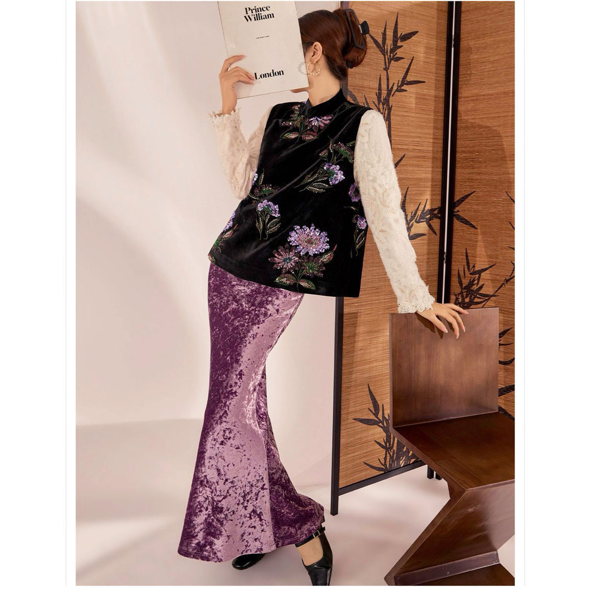 Woman in a black floral top and purple skirt holding a brown bag against a neutral background