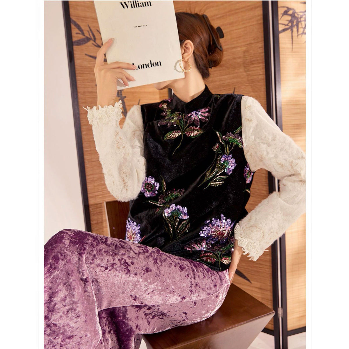 Person wearing a black floral top and purple pants with a white card in front of their face.