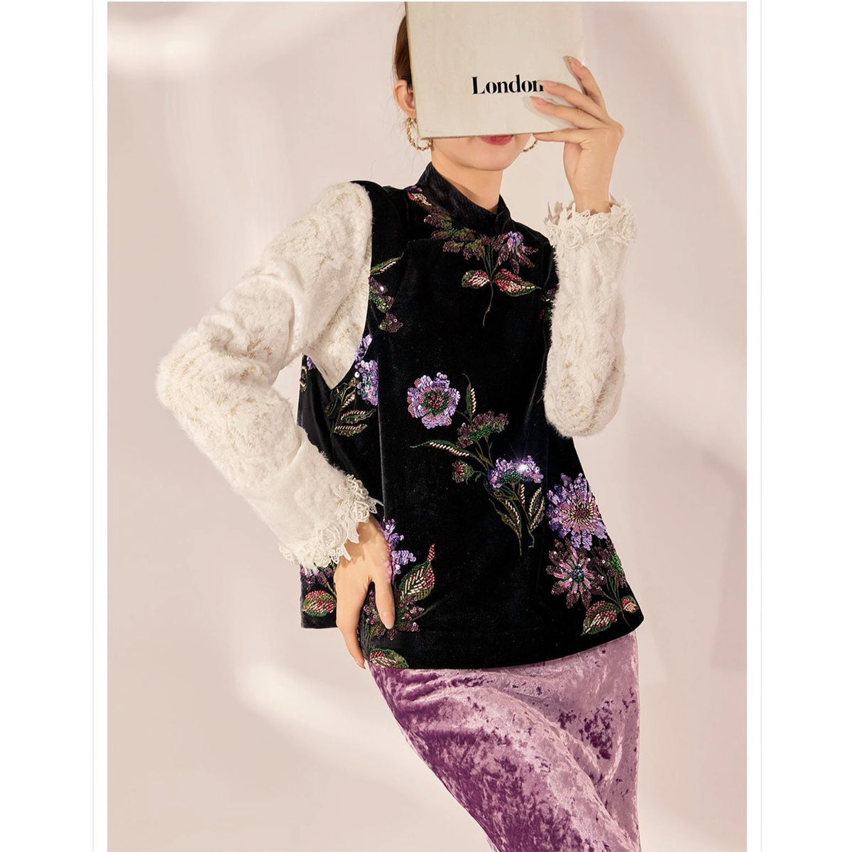 Person wearing a black floral embroidered top with a white lace sleeve and purple skirt, holding a book titled 'London'.