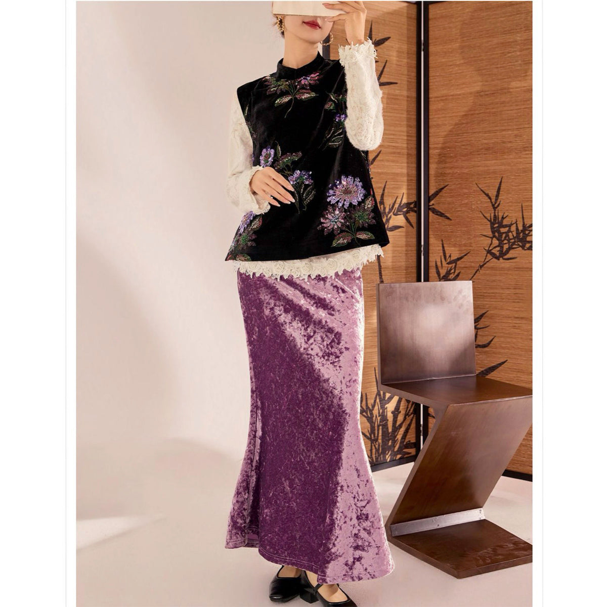 Person wearing a black floral top and purple velvet skirt in a minimalistic setting.
