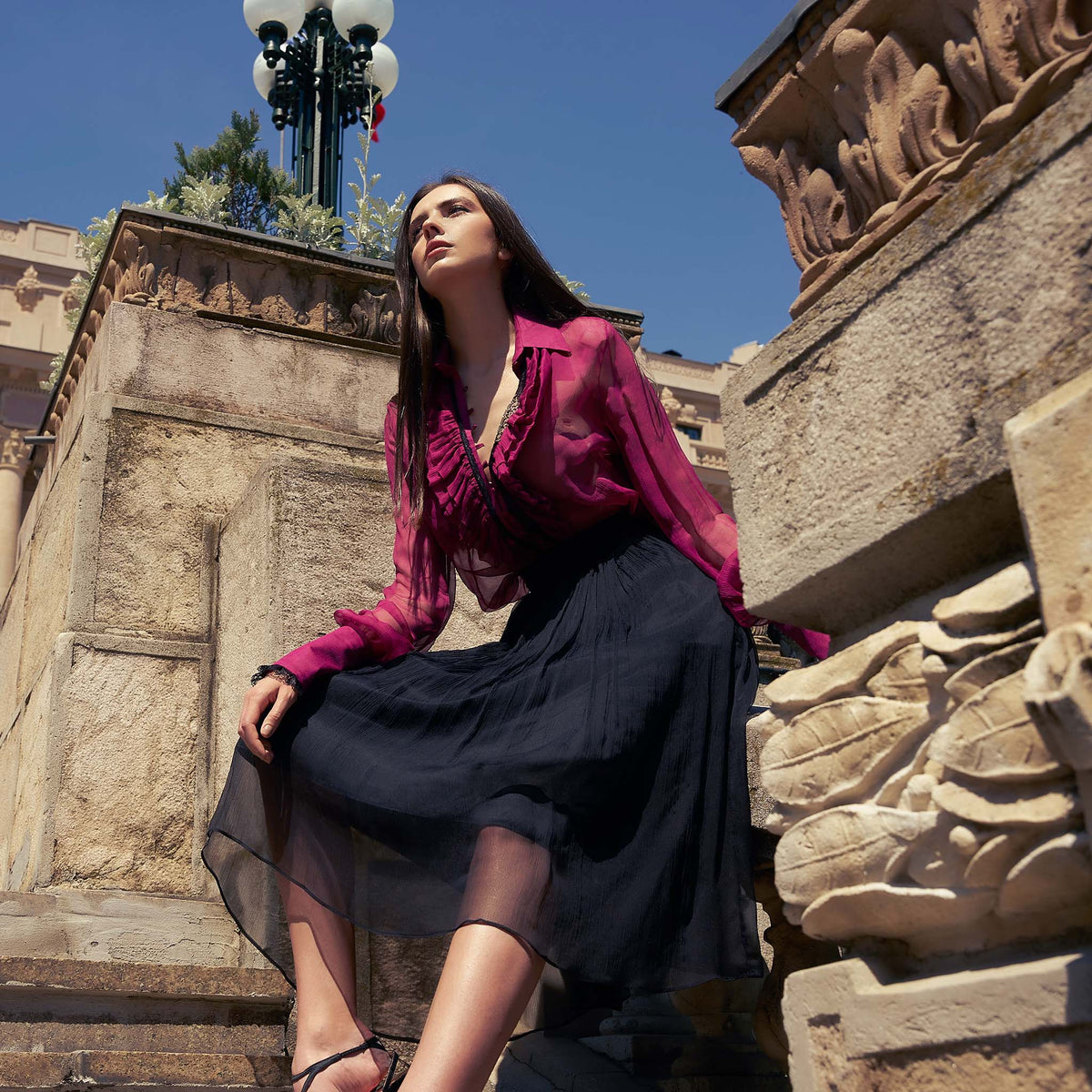 Woman in a pink blouse and black skirt sitting on stone steps with classical architecture.