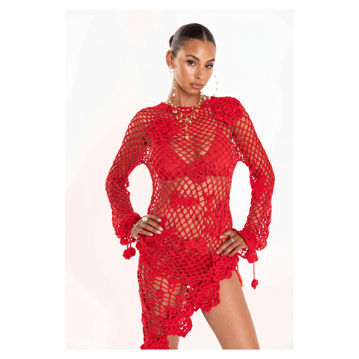 A woman stands confidently in the ANDREEVA Cherry Handmade Crochet Top, featuring bold red color, floral details, and a see-through netted design. Her hair is slicked back in a bun against a plain white background.