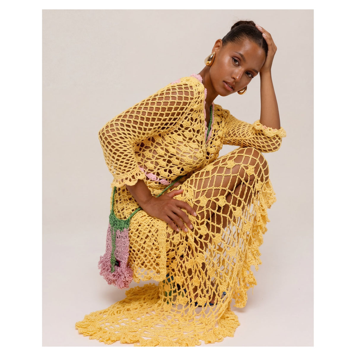 A woman squats, resting one hand on her head, wearing the ANDREEVA California Poppy Yellow Handmade Knit Midi Skirt against a light, plain background.