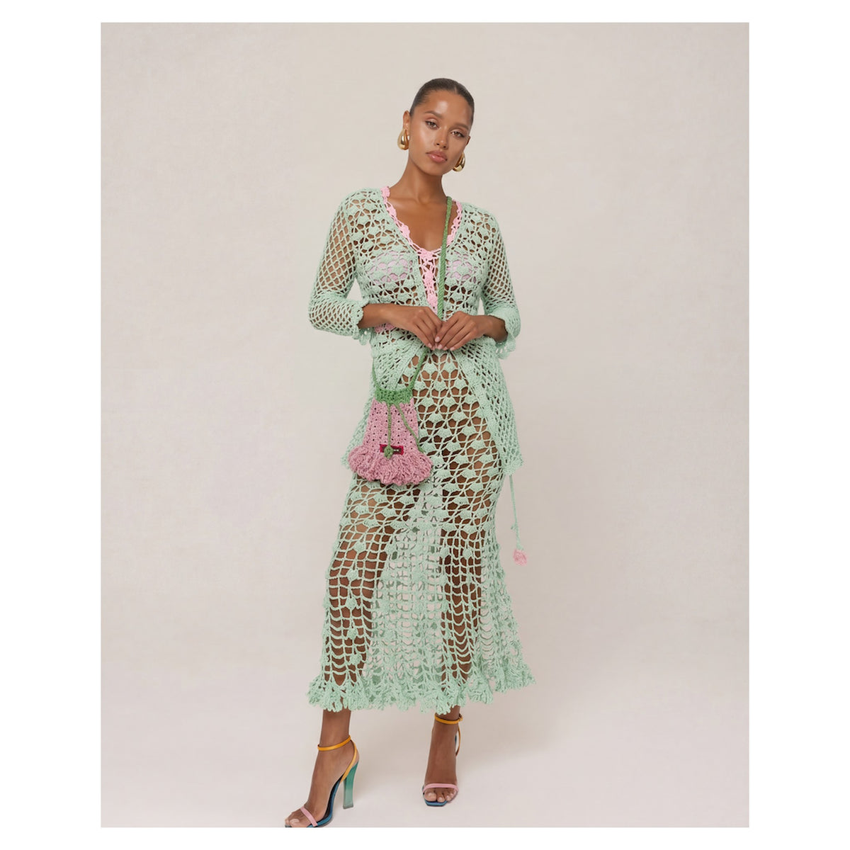 A woman models the ANDREEVA California Mint Handmade Knit Midi Skirt over a pink bikini, accessorized with a pink beaded purse, colorful heels, and hoop earrings, creating chic boho beachwear against a simple light background.