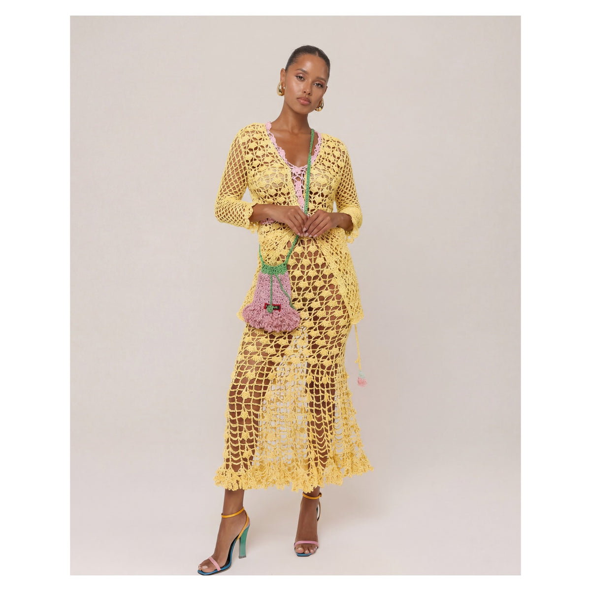 A woman stands against a plain background wearing the ANDREEVA California Yellow Handmade Crochet Shirt over a pink bikini, holding a pink woven handbag with green handles, and pairing it with multicolored high-heeled sandals for boho charm.