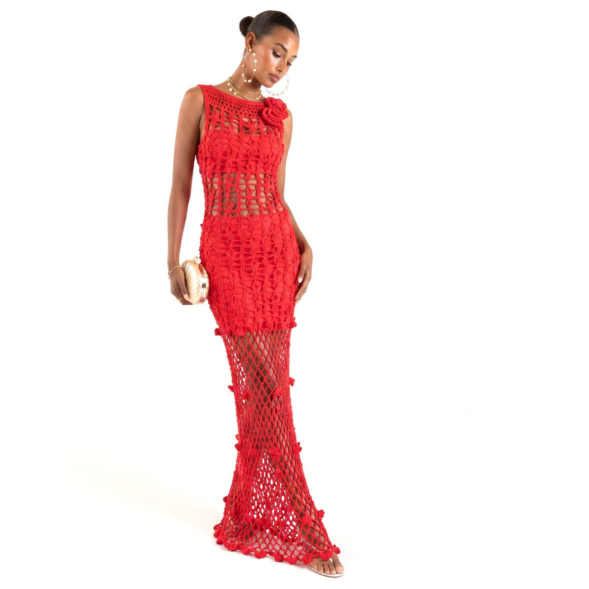 A woman models the ANDREEVA Cherry Handmade Maxi Crochet Dress, a sleeveless floor-length piece with sheer panels and pearl accents. She holds a gold clutch, wears hoop earrings, has her hair in a bun, and stands before a white background.