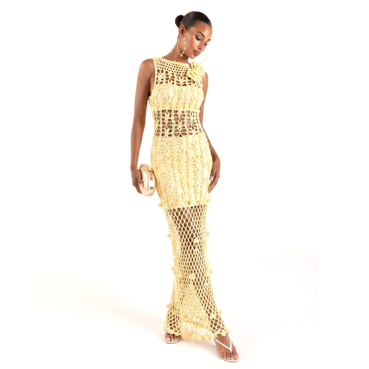 A woman poses in the Yellow Cherry Handmade Maxi Crochet Dress by ANDREEVA, a sleeveless, floor-length crochet dress with floral details and open back. She accessorizes with a small round clutch and strappy heels, her hair styled in a sleek updo.